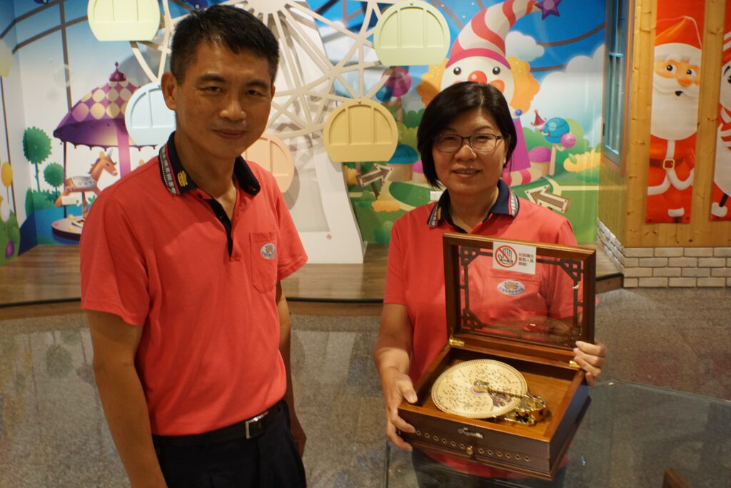 林麗玫經理手持協櫻設計的發條唱盤式音樂盒，外觀由她設計，機芯由左側的張錦財廠長負責。