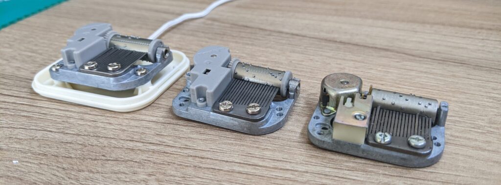 三款迷你機芯並列展示，從右至左依序為 Sankyo 原始設計、協櫻改良版，以及組裝於塑膠殼內的拉繩音樂鈴。