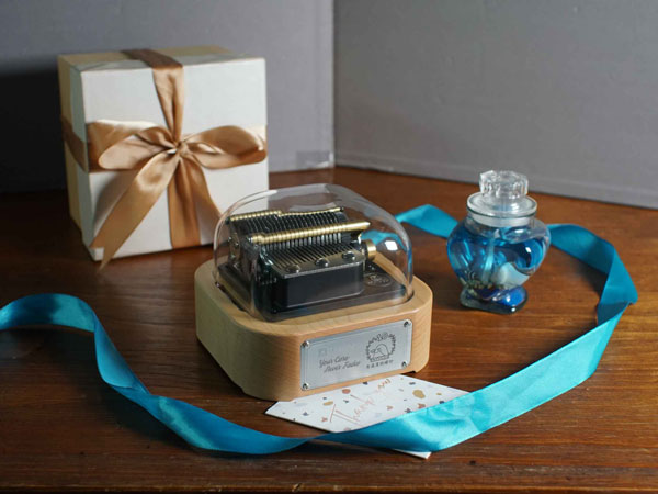 The Muro Box N20 programmable music box with a custom engraved metal nameplate featuring “Your Care Never Fades” and “A Cherished Bond Filled with Warmth”, along with a Japanese White-eye illustration. Beside it are a gold ribboned gift box and a blue diffuser bottle.