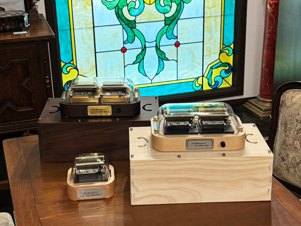 On a wooden table inside the Arashiyama Music Box Museum shop in Kyoto, Japan, the full lineup of Muro Box programmable music boxes is displayed: the Muro Box-N20 Standard Edition, the Muro Box-N40 Standard Edition, the Muro Box-N40 Sublime Edition, and both N40 models paired with their respective N40 Resonance Boxes. The stained glass window in the background adds an artistic ambiance.